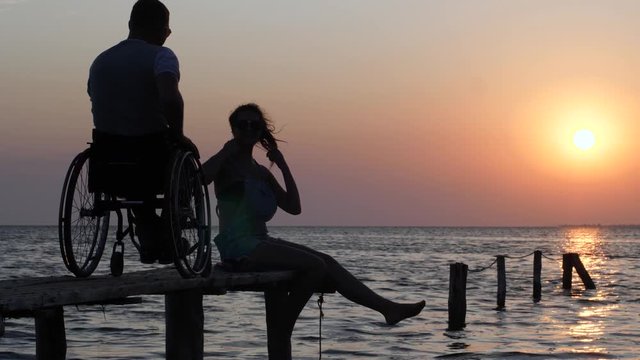 disabled person living a full life, laughing girl in sunglasses sitting on wooden pier and talking with man disabled in wheelchair backdrop of sunset and sea water