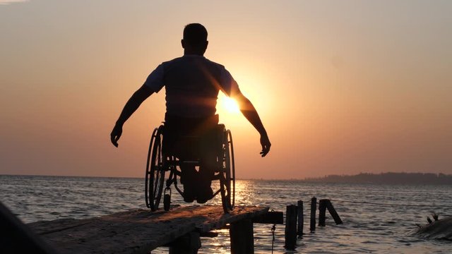 ill male in wheel chair resting and raises his arms backdrop of orange skyline near water on river on summer travel