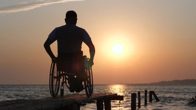 handicapped male on wheel chair resting to jetty on background of orange heaven and water at sundown at summer holiday