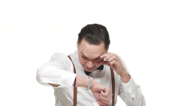 Portrait of artistic mature man wearing white shirt and bow tie looking at his wrist watch with fun, over white background in studio. Concept of emotions