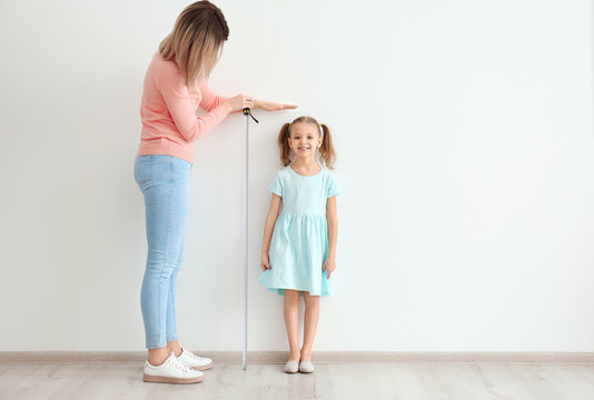 Mother measuring height of little girl near light wall
