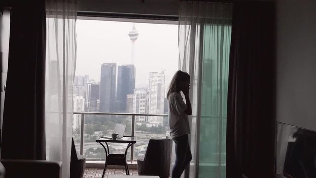 Young female freelancer or businessman ceo talking on the phone, negotiating the panoramic windows and terrace in luxury apartment on business trip. Silhouette of woman using smartphone.
