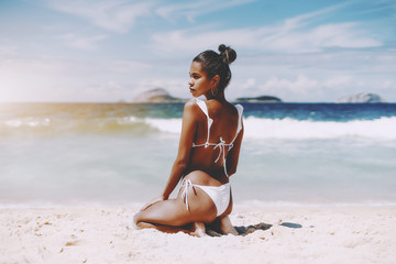 Hottie young African American female model is sitting on the sand half-turned to camera in front of ocean; rear view of foxy Brazilian girl sitting on the beach of warm summer sea with horizon behind - Powered by Adobe