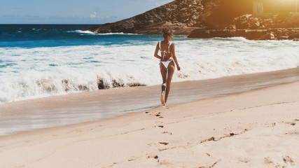 Dainty thin young Brazilian female is going away into the distance of sandy beach to the rocks in front; sexy black girl in swimsuit is walking on the ocean beach leaving footprints in the sand - obrazy, fototapety, plakaty
