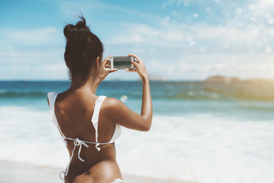 View from behind of svelte young Brazilian female in swimsuit after swimming in ocean and now she is taking pictures of ocean and islands in the defocused background using camera of her smartphone
