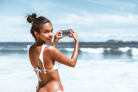Rear view of svelte young Brazilian girl in swimsuit with wet skin, half-turned and looking at camera while taking photos of beautiful seascape and islands in front of her after swimming in ocean