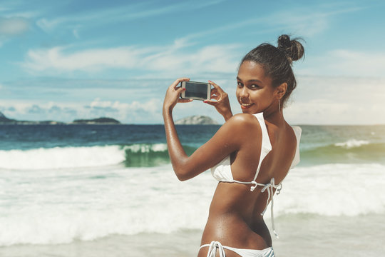 View from behind of sexy young black girl in swimsuit with wet skin, half-turned and looking at camera with smile while taking photos of ocean and horizon using her smartphone on sunny summer day