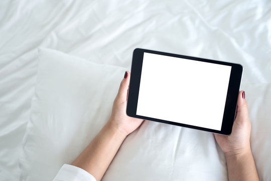 Top view mockup image of a woman holding black tablet pc with blank desktop white screen while sitting on a white bed background