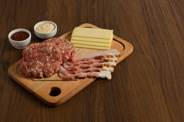 Ingredients for cooking burgers. Raw ground beef meat cutlets on wooden chopping board, cheese, bacon,  over wooden background