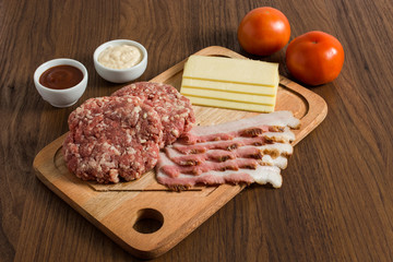 Ingredients for cooking burgers. Raw ground beef meat cutlets on wooden chopping board, cheese, bacon,  over wooden background