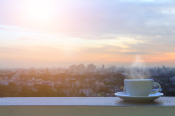 White coffee cup and softy smoke put on sky terrace border with blurred morning city scape and sunrise effect, Selective focus and copy space