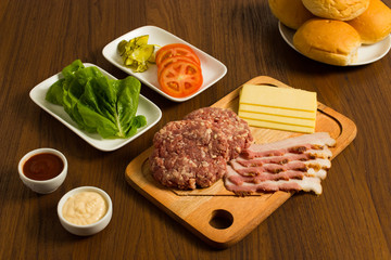 Ingredients for cooking burgers. Raw ground beef meat cutlets on wooden chopping board, tomatoes, greens, pickles, ketchup, cheese,  over wooden background 