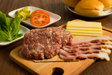 Ingredients for cooking burgers. Raw ground beef meat cutlets on wooden chopping board, tomatoes, greens, pickles, ketchup, cheese,  over wooden background 