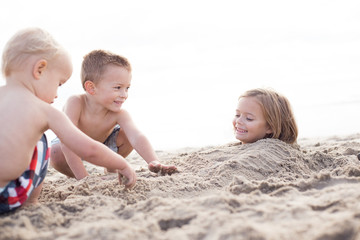 Two little boys buryind a smiling girl in the sand on the beach