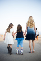 Two older sisters helping younger sister rollerskate on a sidewalk by the beach