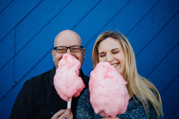 A man and a woman smiling and holding sticky, pink cotton candy infront of a blue wall