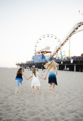 Three young girls running in the sand towards the Santa Monica Pier ferris wheel and rollercoaster