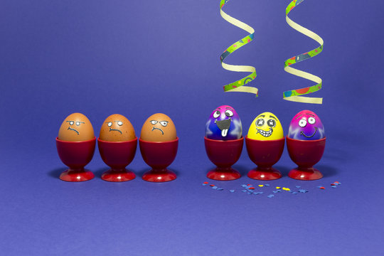 Group of colorful painted Easter eggs with funny cartoon style faces, confetti and paper streamers and group of grumpy looking brown eggs in red plastic egg cups on purple background