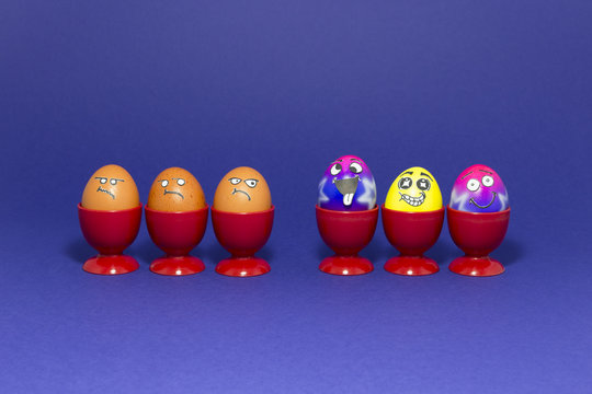 Group of colorful painted Easter eggs with funny cartoon style faces and group of grumpy looking brown eggs in red plastic egg cups on purple background
