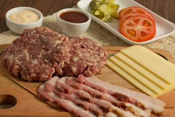 Ingredients for cooking burgers. Raw ground beef meat cutlets on wooden chopping board, tomatoes, greens, pickles, ketchup, cheese,  over wooden background 