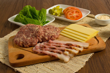 Ingredients for cooking burgers. Raw ground beef meat cutlets on wooden chopping board, tomatoes, greens, pickles, ketchup, cheese,  over wooden background 