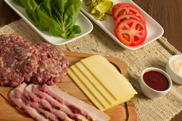 Ingredients for cooking burgers. Raw ground beef meat cutlets on wooden chopping board, tomatoes, greens, pickles, ketchup, cheese,  over wooden background 