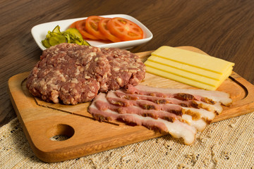 Ingredients for cooking burgers. Raw ground beef meat cutlets on wooden chopping board, tomatoes, greens, pickles, ketchup, cheese,  over wooden background 