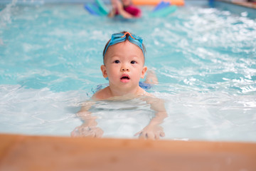 Fototapeta na wymiar Cute little Asian 18 months / 1 year old toddler boy child in trunks wear swimming goggles learn to swim at indoor pool, Swimming school for small children. Happy kid enjoy active lifestyle concept