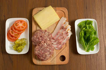 Ingredients for cooking burgers. Raw ground beef meat cutlets on wooden chopping board, tomatoes, greens, pickles, ketchup, cheese,  over wooden background - Top View