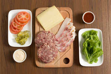 Ingredients for cooking burgers. Raw ground beef meat cutlets on wooden chopping board, tomatoes, greens, pickles, ketchup, cheese,  over wooden background - Top View