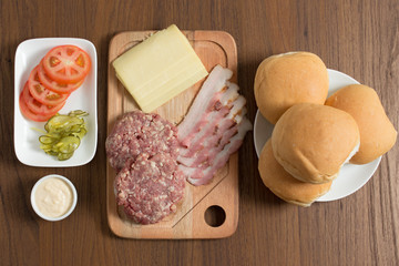 Ingredients for cooking burgers. Raw ground beef meat cutlets on wooden chopping board, tomatoes, greens, pickles, ketchup, cheese,  over wooden background - Top View