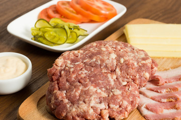 Ingredients for cooking burgers. Raw ground beef meat cutlets on wooden chopping board, tomatoes, greens, pickles, ketchup, cheese,  over wooden background - Top View