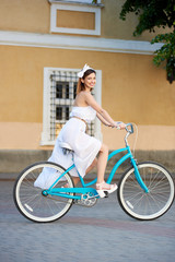 Fototapeta premium Attractive young female in a white dress is looking to the camera while riding a blue vintage bike down beautiful city street on a summer day. Pretty woman high heels turquoise bicycle green tree.
