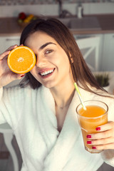 Beautiful girl wears white bathrobe with glass of orange juice and fruit, kitchen morning sunset view