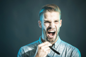 Screaming unshaven macho in shirt with straight razor near his beard. Young man with beard and...