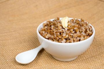 cooked buckwheat heap with butter in white bowl with spoon over sackcloth