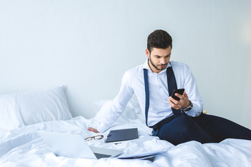 businessman working with smartphone, documents and laptop on bed in morning