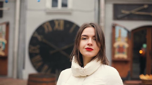 Brunette pretty young women portrait with red lips smiling, flirting, touches hair and looking in to the camera. Old city background. Cold season. Slow motion.