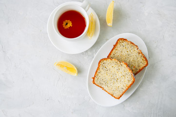 Slices of homemade lemon poppy seed cake served on a white plate, a cup of tea on a white stone background.