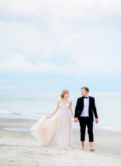 Bride and groom hold each other hands tender walking along the beach by the sea