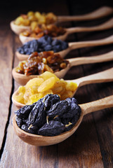 Black, brown and yellow raisins in spoons on a dark wooden background.