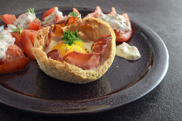 muffin from egg and bacon in toasted bread, tomatoes and cheese cream with herbs on a dark plate on rustic slate, creative breakfast, brunch or snack