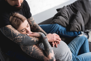 cropped image of tattooed boyfriend hugging pregnant girlfriend on sofa at home