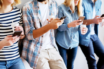 Group of young modern people holding mobiles and coffee in the paper cup leaning against the wall while waiting or having a break. Modern mobile obsession.