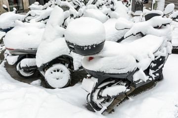 Snow-covered two and three-wheel scooters parked outdoors in the streets of Paris.