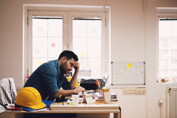 Very sleepy and tired young modern engineer entrepreneur looking at reports on a laptop in his office.