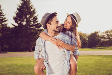 Portrait of cute romantic bearded brunet guy in checkered shirt, dreamy lady rides him on rear. Leisure, chill happiness, lawn stroll, relax, romance lifestyle, well dressed partners posing © deagreez