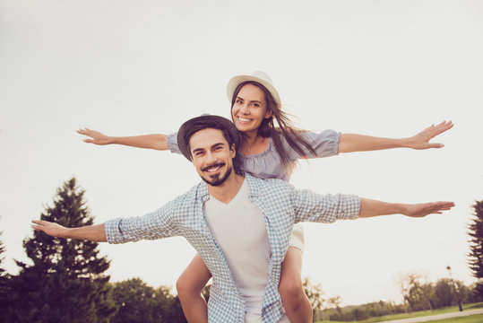 Cute romantic bearded brunet in checkered shirt, dreamy lady rides him on rear. Leisure, chill happiness, lawn stroll, relax, romance lifestyle, well dressed in blue, partners posing in headwear