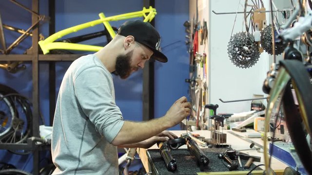 A man bike mechanic with a beard assembles a mountain bike in his workshop.
