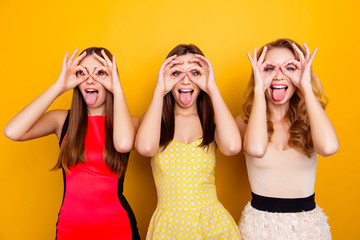 Three pretty, charming, attractive, nice, trendy, crazy girls showing binoculars with fingers, hands and gesture tongue out, foolish over yellow background